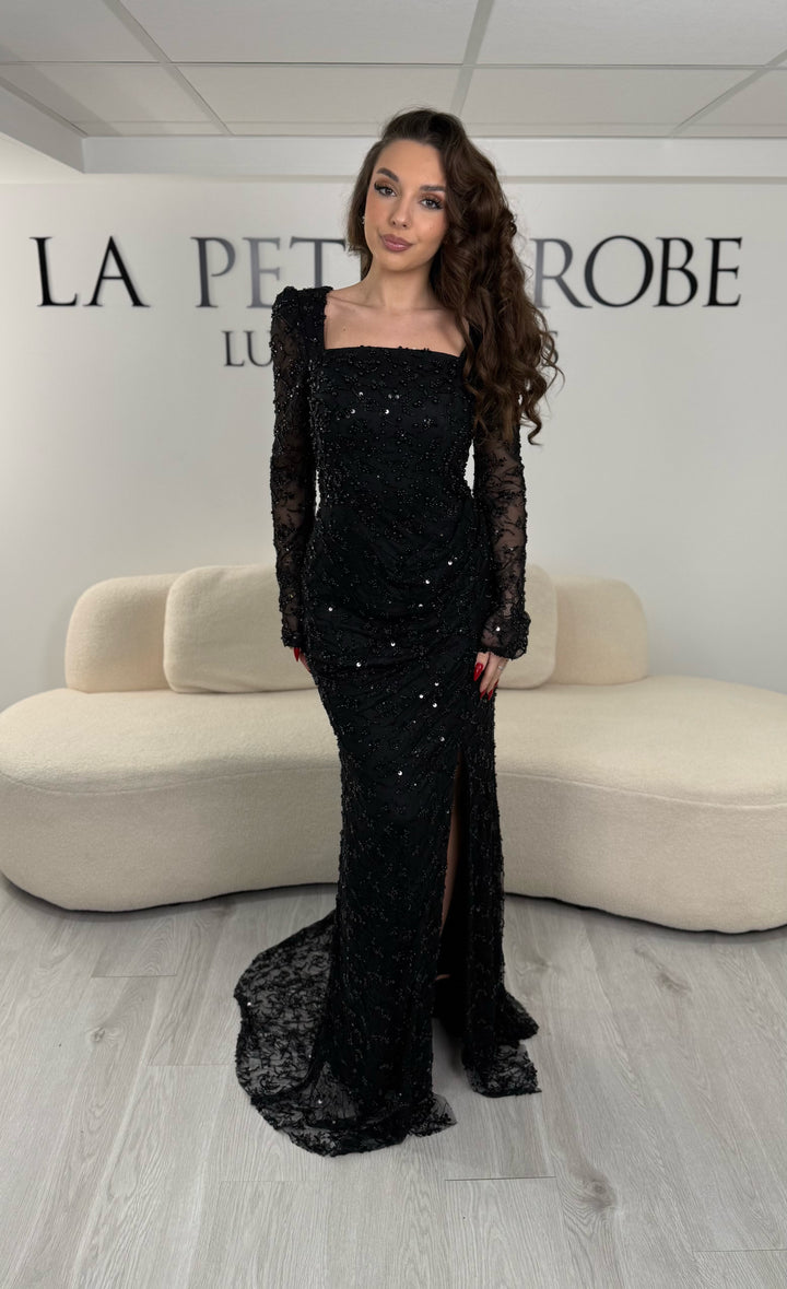 Robe de soirée noire élégante avec col carré, manches longues et fente glamour. Idéale pour un look chic, lumineux et raffiné.