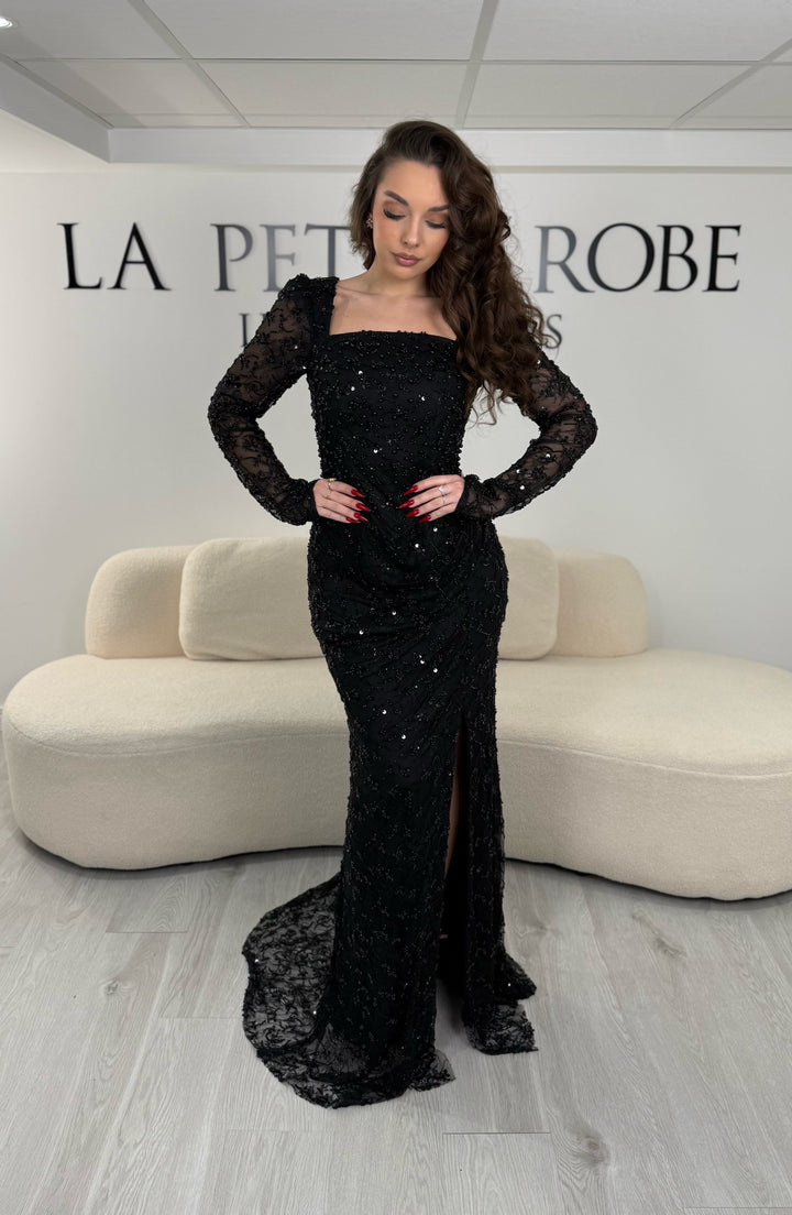 Robe de soirée noire élégante avec col carré, manches longues et fente glamour. Idéale pour un look chic, lumineux et raffiné.