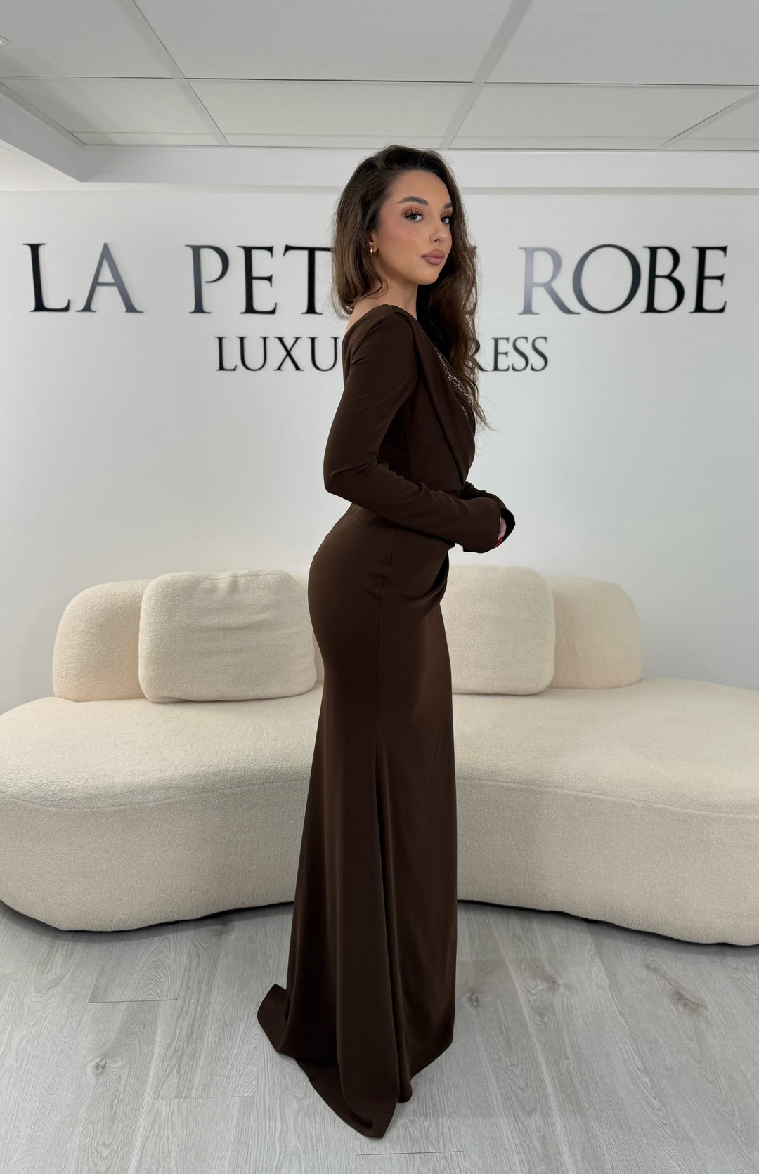 Robe de soirée marron avec col asymétrique et manches longues. Coupe ajustée avec fente sur le côté, idéale pour une allure élégante et sophistiquée.