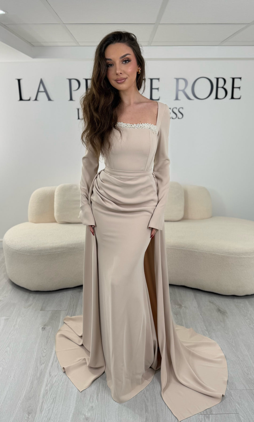 Robe de soirée beige avec col carré et manches longues. Coupe ajustée sans fente avec traîne amovible, idéale pour une allure élégante et raffinée.