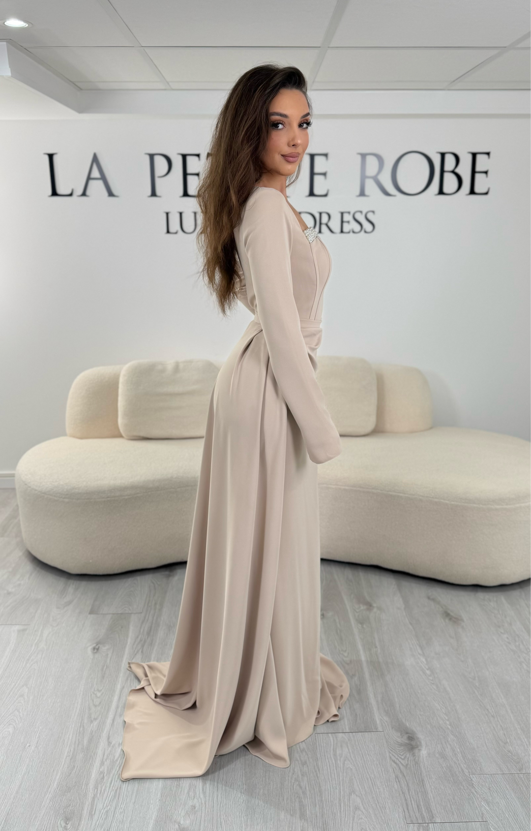 Robe de soirée beige avec col carré et manches longues. Coupe ajustée sans fente avec traîne amovible, idéale pour une allure élégante et raffinée.
