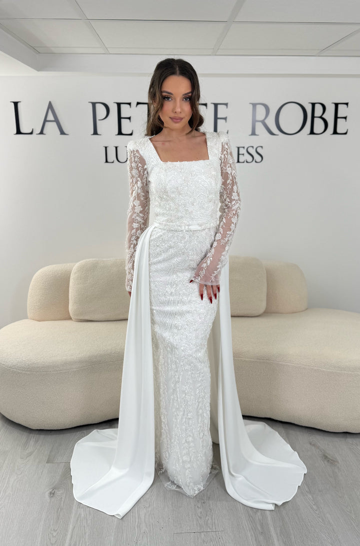 Robe de soirée blanche avec col carré et manches longues brodées. Coupe ajustée sans fente avec traîne amovible, idéale pour une allure élégante.