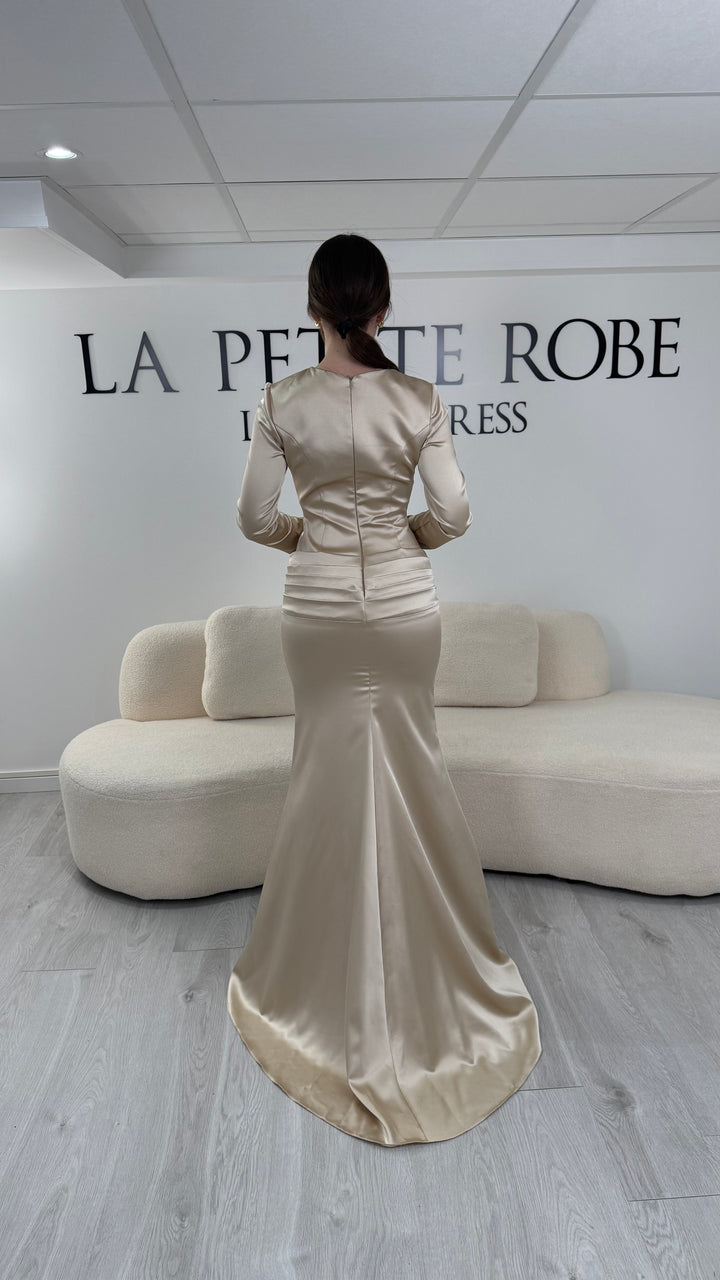 Robe de soirée beige avec manches longues et coupe ajustée. Sans fente, tissu satiné et détails élégants pour une allure sophistiquée.