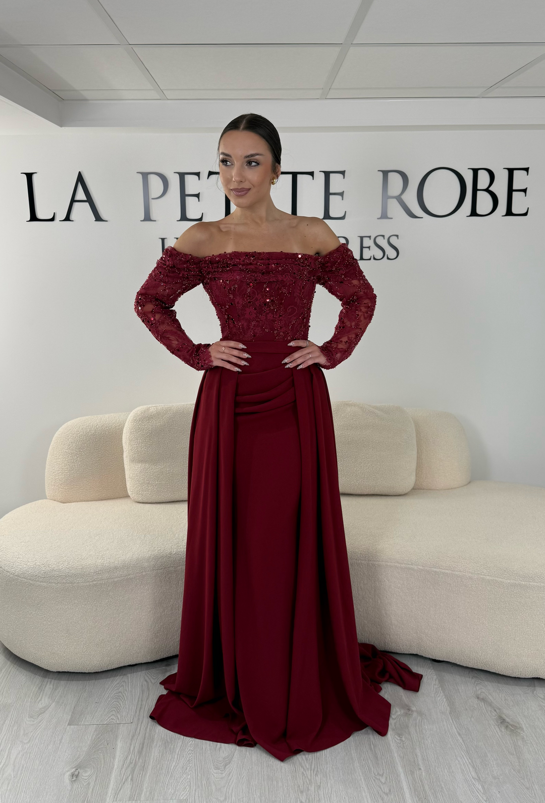Robe de soirée bordeaux longue avec dentelle raffinée, manches longues et décolleté bardot. Modèle élégant sans fente avec traîne amovible, idéal pour mariage et cérémonie.