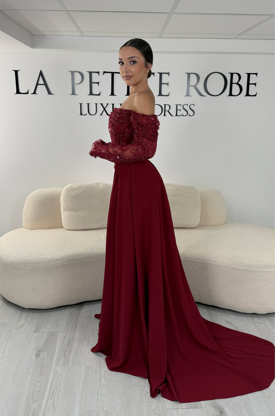 Robe de soirée bordeaux longue avec dentelle raffinée, manches longues et décolleté bardot. Modèle élégant sans fente avec traîne amovible, idéal pour mariage et cérémonie.