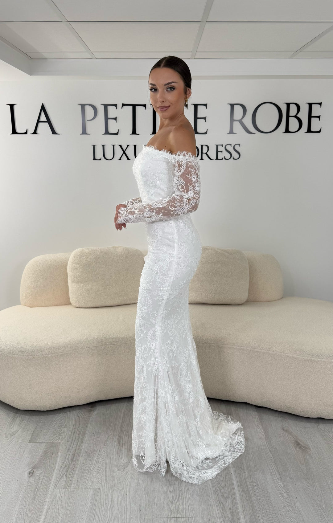 Robe de mariée blanche longue en dentelle et sequins avec col bardot et manches longues. Une robe élégante sans fente idéale pour cérémonie, mariage civil et réception.