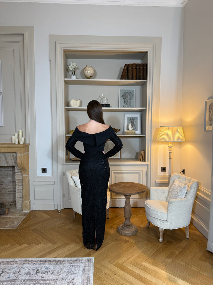 Robe de soirée noire élégante avec col bateau, manches longues et fente à l’arrière. Une robe chic et intemporelle pour vos événements.