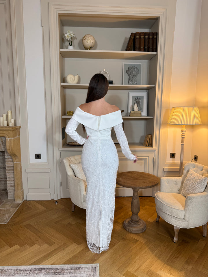 Robe de cérémonie blanche élégante avec col bateau, manches longues et fente à l’arrière. Une robe raffinée pour vos événements.