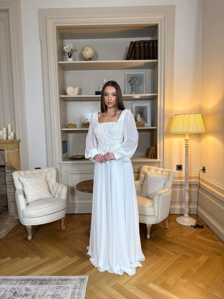 Longue robe de mariée blanche élégante à manches longues, col carré et coupe fluide sans fente. Une robe raffinée pour un mariage chic.