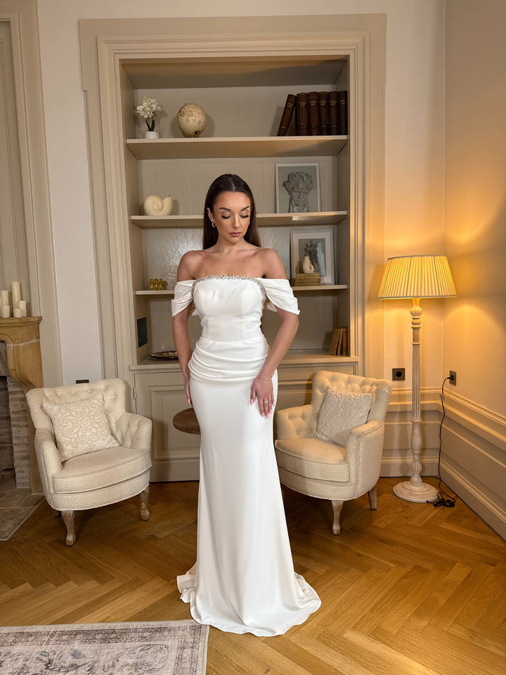 Robe de mariée blanche élégante au col bateau, coupe fluide sans fente. Une robe raffinée et intemporelle pour un mariage chic.