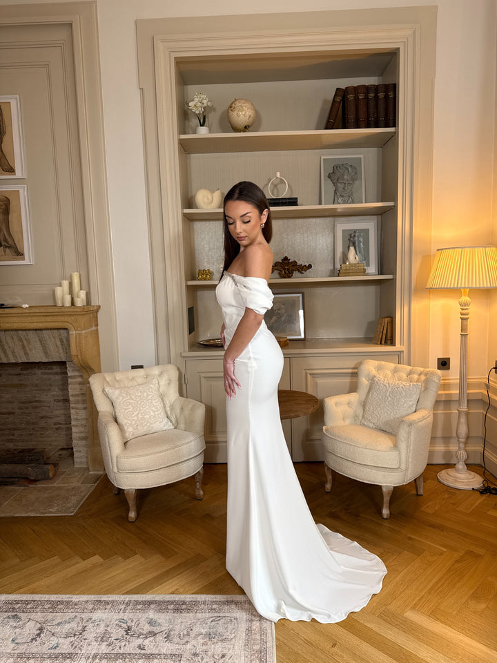 Robe de mariée blanche élégante au col bateau, coupe fluide sans fente. Une robe raffinée et intemporelle pour un mariage chic.