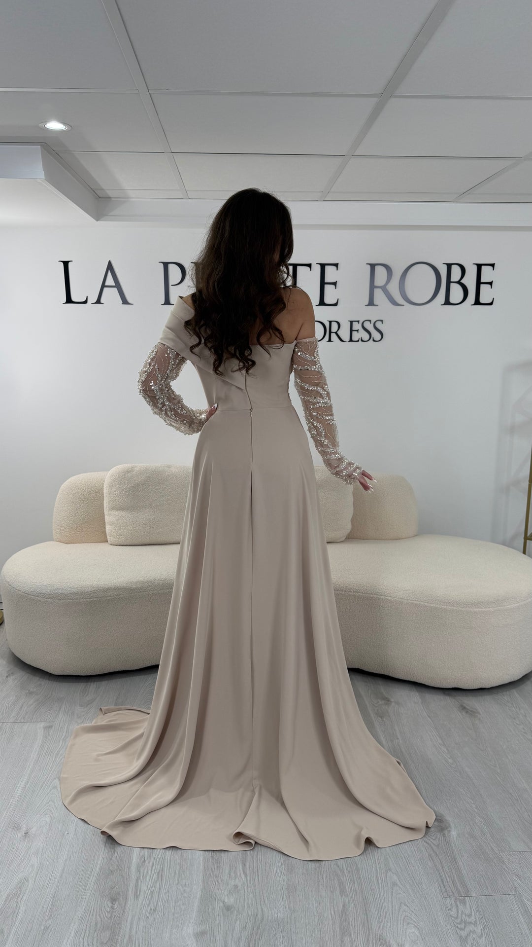 Longue robe de soirée beige chic avec col asymétrique, manches longues transparentes et fente latérale élégante. Coupe fluide et raffinée idéale pour mariage, gala et réception.