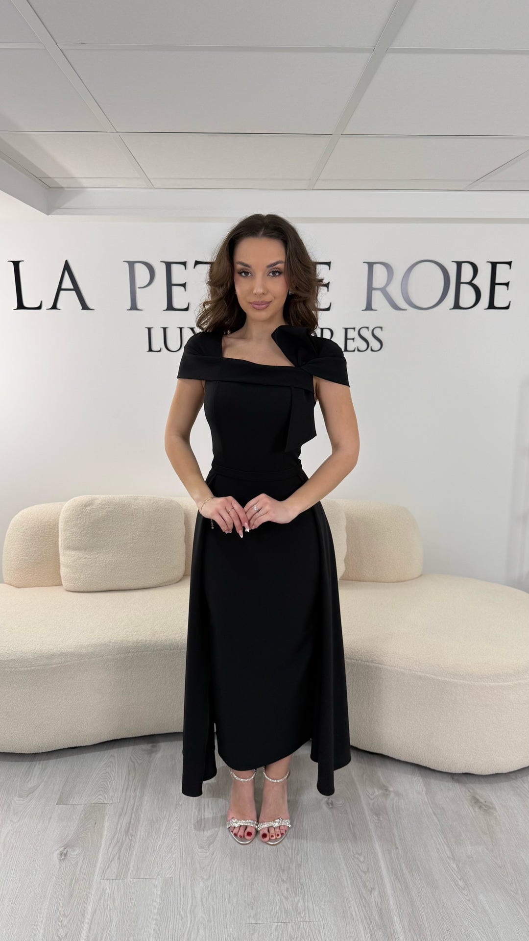 Robe de soirée noire midi avec col bateau élégant et coupe sans manche. Sans fente, silhouette raffinée idéale pour réception et soirée chic.