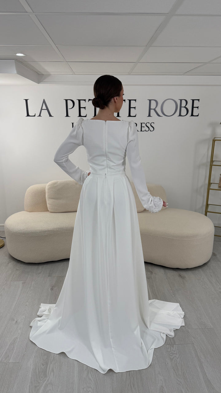 Robe de mariée blanche élégante avec col rond et manches longues. Robe longue sans fente, coupe raffinée et détails perlés, parfaite pour une cérémonie de mariage chic.