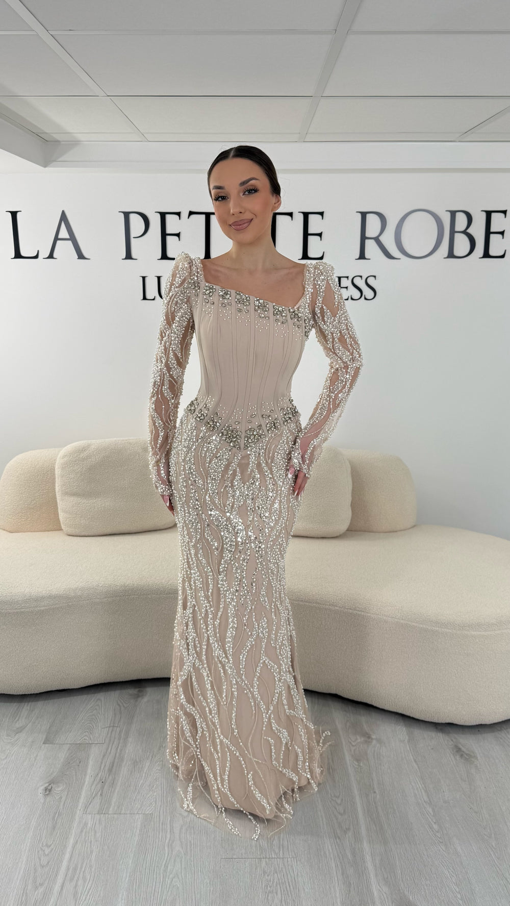 Robe de soirée beige élégante avec col asymétrique et manches longues brodées. Robe longue sans fente idéale pour gala ou soirée chic.