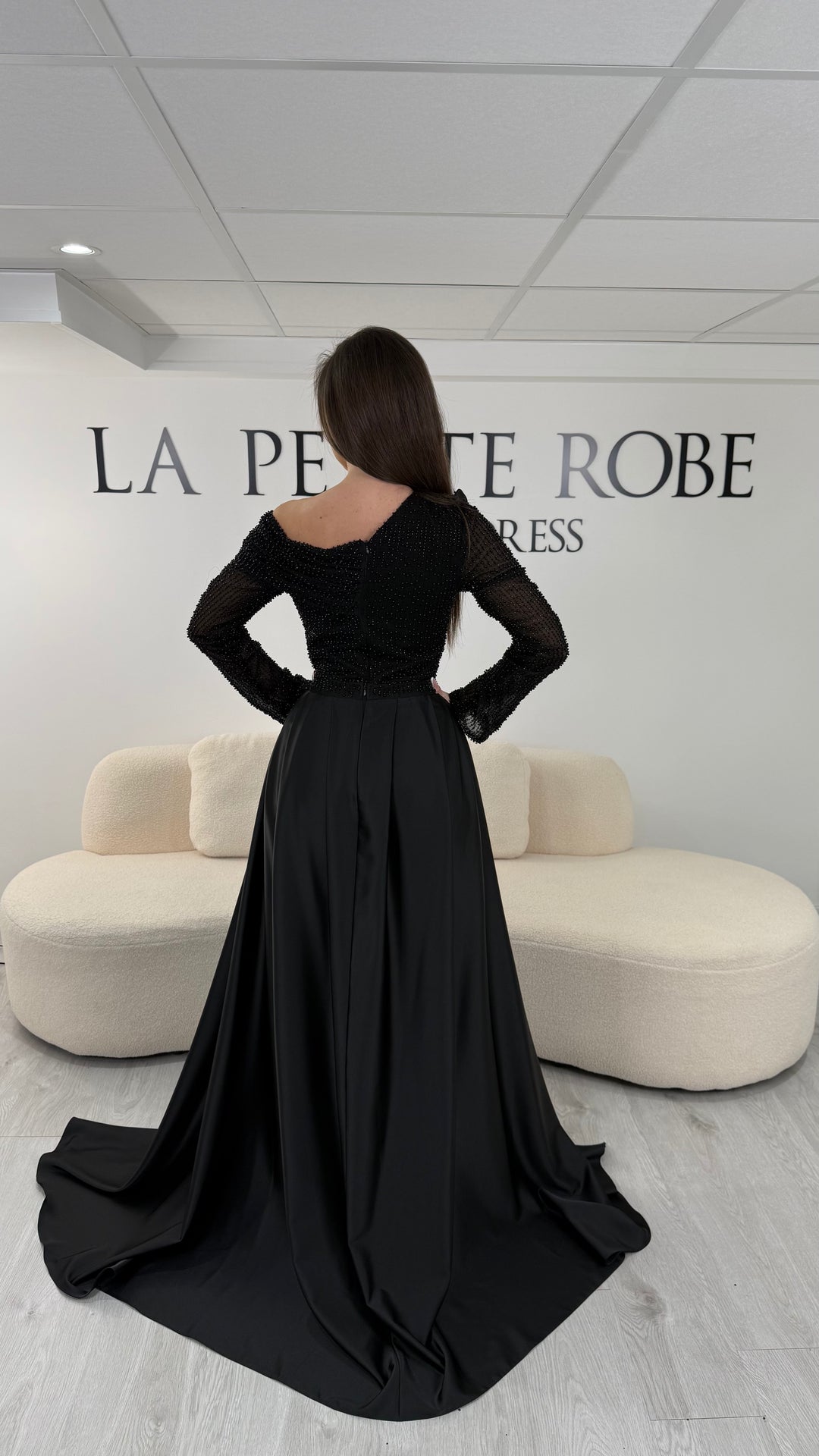 Robe de soirée noire longue, sans fente, avec col asymétrique, manches longues et surjupe amovible. Élégante et modulable.