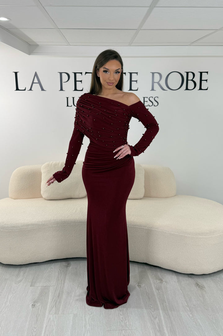 Robe de soirée bordeaux longue, sans fente, avec col asymétrique et manches longues. Élégante avec détails perlés pour vos soirées.